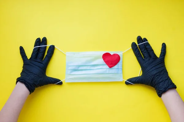Doctor holds protective face mask with heart in hands in black nitril gloves on yellow background. Pandemic insurance, airborne diseases, Grippe. Concept care and treatment with love