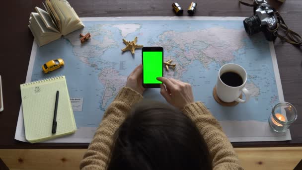 Bovenaanzicht. Kaart van de wereld op de tafel, een vrouw die het plannen van een vakantie. Womens handen gebruiken een smartphone met groen scherm — Stockvideo