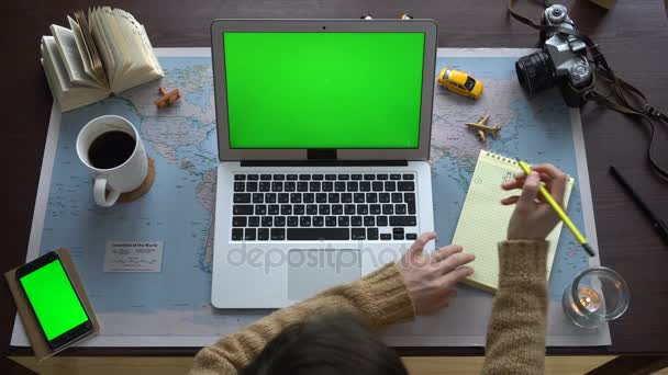 Vista superior. Mapa do mundo na mesa, uma mulher a planear umas férias. Um laptop e um smartphone com tela verde — Vídeo de Stock