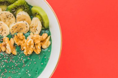 Kırmızı zemin üzerinde muz, ceviz, kivi ve chia tohumları olan bir spirulina smoothie kasesi.