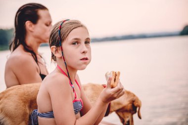 Annesi ve köpek ile nehir kenarında oturan ve sandviç yeme küçük kız