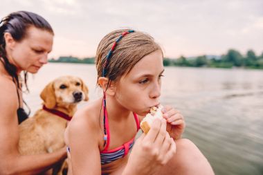 Annesi ve köpek ile nehir kenarında oturan ve sandviç yeme küçük kız