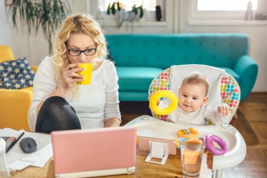 Kahve içme ve bebeğini akıllı telefon üzerinde çizgi film izlerken sırasında dizüstü bilgisayar üzerinde çalışan bir masada oturan gözlük takmış anne