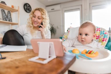 Bebek anne akıllı telefonda konuşurken ve notlar aşağı evde ofis yazma sırasında akıllı telefon izlerken
