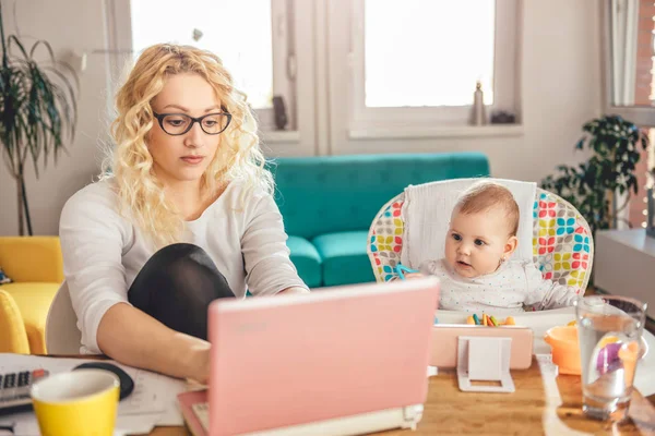 Mother wearing eyeglasses working at home office on laptop and taking care of her baby