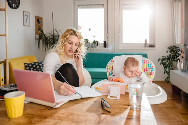 Mother talking on smart phone and writing down notes at home office and taking care of her baby