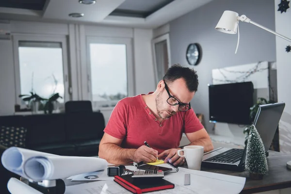 Man writing notes on memo pad at busy desk in home office