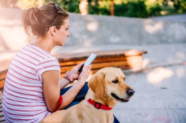 Parkta köpeğiyle oturan ve telefon kullanan bir kadın.