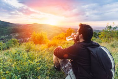 Man with beard drinking water from plastic bottle and enjoying mountain sunset. clipart