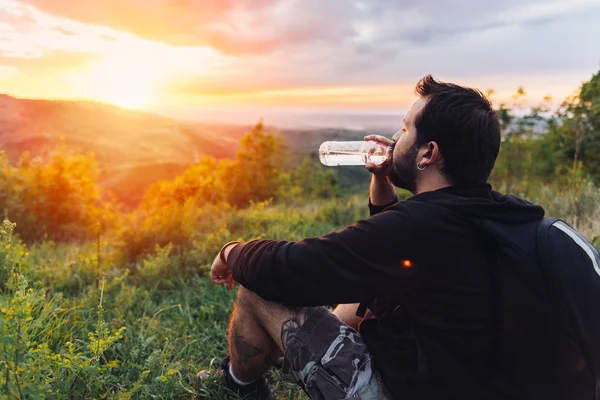 Man with beard drinking water from plastic bottle and enjoying mountain sunset.