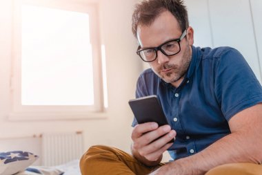 Yatakta oturmuş ve akıllı telefon kullanarak mavi gömlekli gözlüklü adam.