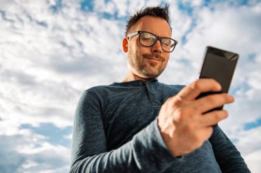 Akıllı telefon açık kullanarak gözlüklü adam. O mavi tişört giyiyor.