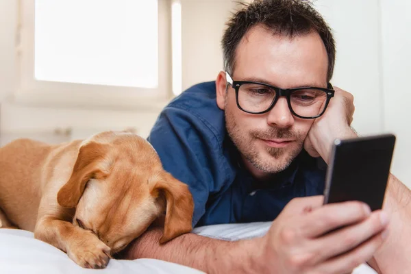 Köpek yatakta döşeme ve akıllı telefon kullanarak mavi gömlekli gözlüklü adam.