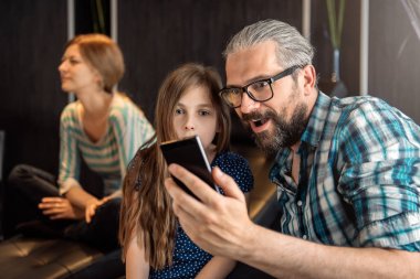 Baba ve kızı ile akıllı telefon, arkalarındaki haber sürpriz olduğunu koltukta oturan karısını.