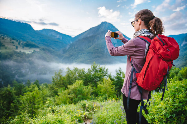 Woman with red backpack hiking on the mountain and taking picture with smart phone