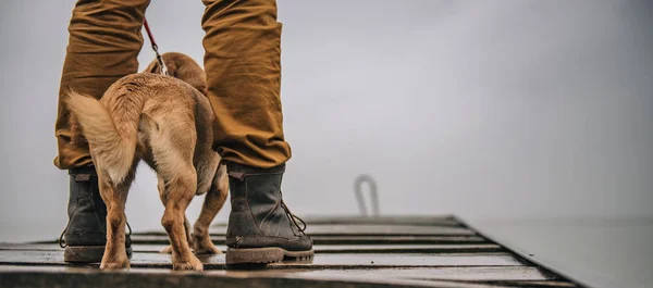 Man in boots protecting his dog from rain and standing on old wooden deck