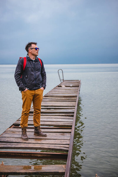 Человек, стоящий на деревянном причале в бурную погоду. На нем красный рюкзак, синяя куртка и кожаные сапоги.
