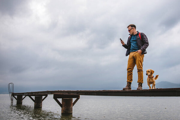 Man standing on wooden dock on stormy weather with his dog and using smart phone. He is wearing red backpack, blue jacket and leather boots