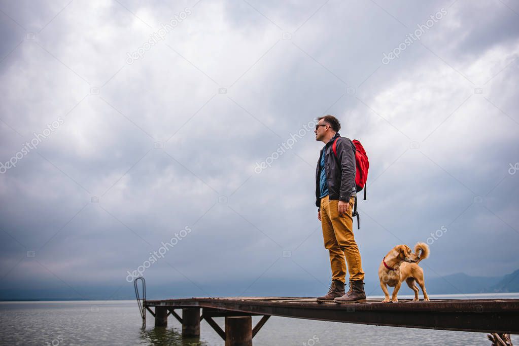 Man standing on wooden dock on stormy weather with his dog. He is wearing red backpack, blue jacket and leather boots