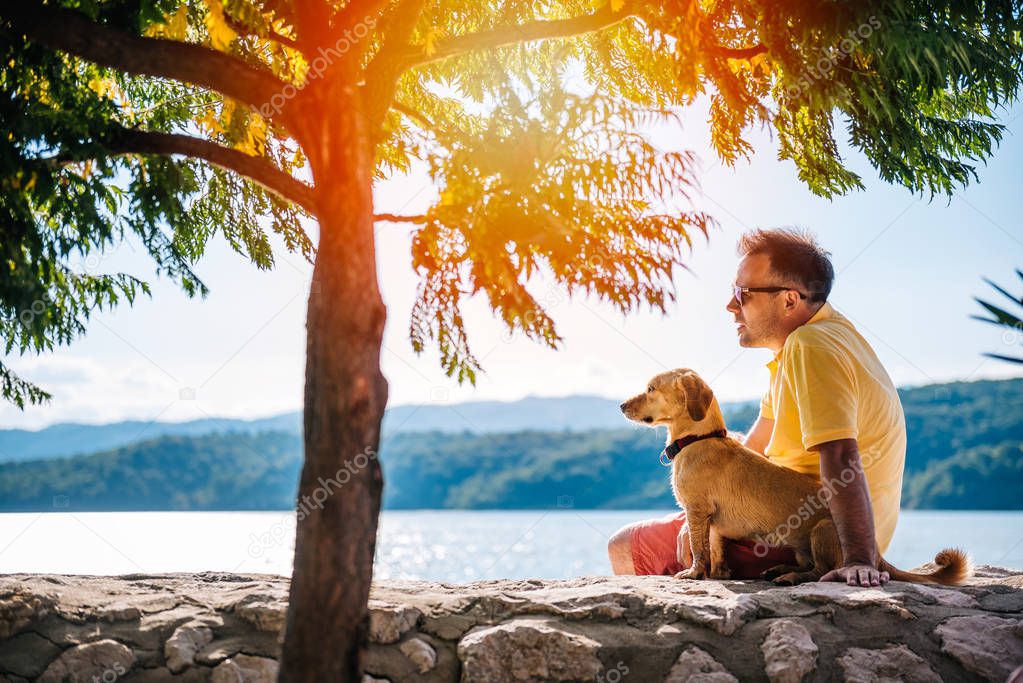 Man in yellow shirt and sunglasses sitting with his dog on a stone wall beside small tree 