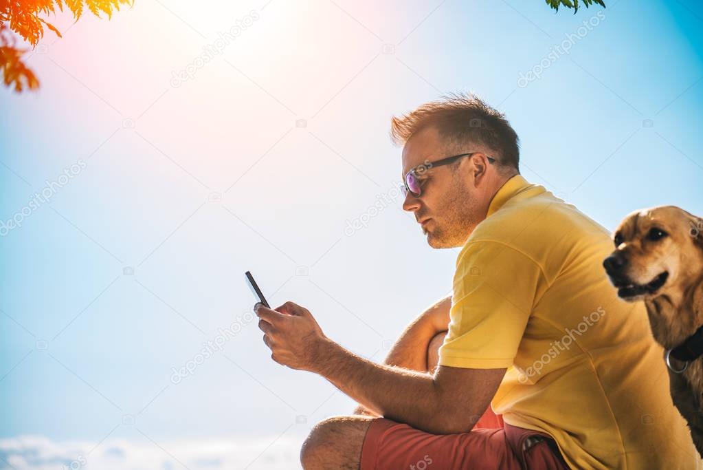 Man in yellow shirt and sunglasses using smart phone, beside him is a small yellow dog
