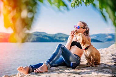 Bununla birlikte güneş gözlüklü siyah bikini ve kahve içme ve küçük sarı köpeği ile sahilde kurabiye yemek kot giyen kadın