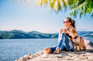 Bununla birlikte güneş gözlüklü siyah bikini ve oturan kot giyen kadın taş küçük sarı köpeğiyle rıhtım.