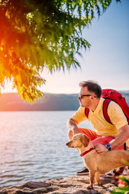 Adam sarı tişört, güneş gözlüğü ve küçük sarı köpeği ile deniz kenarında kırmızı sırt çantası çömelme 