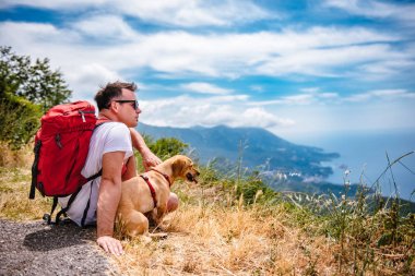 Adam kırmızı sırt çantası ve dağın tepesinde oturan ve denizde seyir ufuk küçük sarı köpek