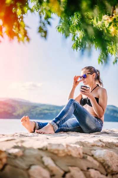 Woman Sunglasses Wearing Black Bikini Jeans Sitting Stone Dock Drinking — Stock Photo, Image