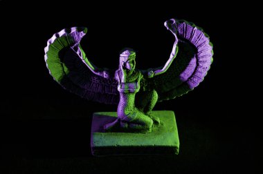 Closeup to a Isis. female old winged egyptian god mini figurine iluminated with green and violet lights over black background clipart