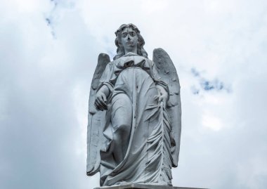Eski bir melek heykeli, anıt mezarlığın tepesine alçak açıyla bakıyor. Arkaplanda mavi ve bulutlu bir gökyüzü