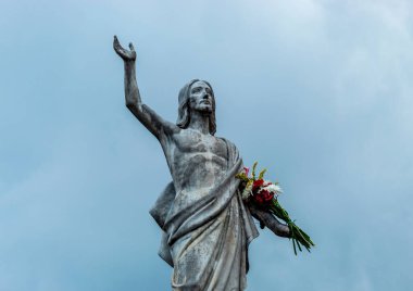 Eski bir İsa heykeli dirildi. Elinde çok renkli bir buket çiçek ve arka planda mavi bulutlu bir gökyüzü vardı.