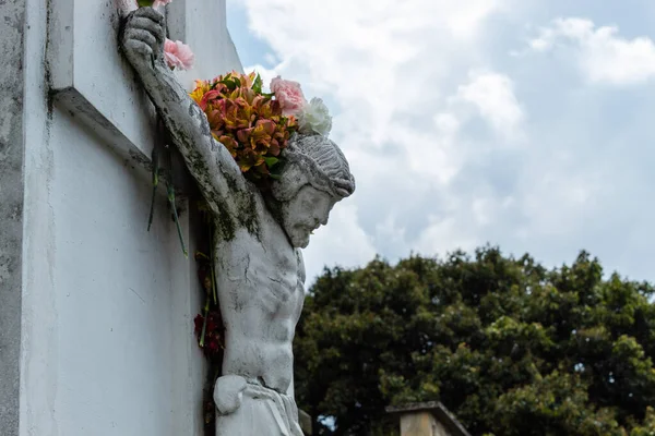 Crucified White Jesus Christ Stone into a Cemetery adorned with multicolor flowers in hands, arms and flowers like a crown in head. Big tree and cloudy sky at background