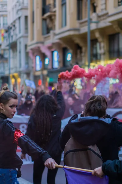 Uluslararası Kadınlar Günü 'nde düzenlenen 8 M yürüyüşleri sırasında arka planda kırmızı işaret fişeği tutan genç bir kadını koruyan menekşe rengi bir bayrak tutan kadınlar zinciri oluşturuyor.