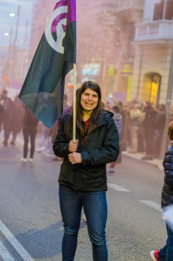 Uluslararası Kadınlar Günü 'nde 8M yürüyüşü sırasında kalabalığın ortasında menekşe siyah bayrak sallayan genç bir kadın haklarını iddia ederek yürür.