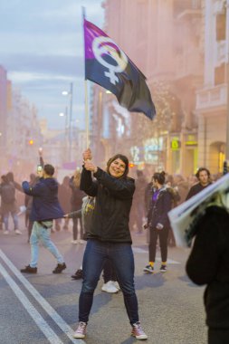 Uluslararası Kadınlar Günü 'nde 8M yürüyüşü sırasında kalabalığın ortasında menekşe siyah bayrak sallayan genç bir kadın haklarını iddia ederek yürür.