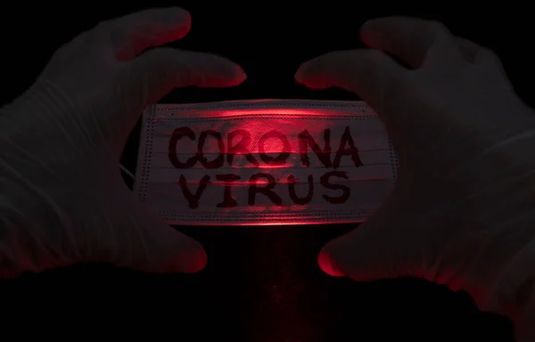 白い滅菌手袋の近くに黒い背景に赤い光に照らされたCoronavirusの赤い碑文で白い顔のマスクに怒りの手の表情を作りますCovid 2019 — ストック写真