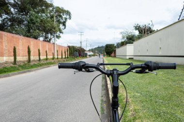 Yeşil bir bahçenin yakınındaki banliyöde asfalt bir yol ve arka planda bulanık dağlar olan siyah bir dağ bisikleti kulpu barına yakın.