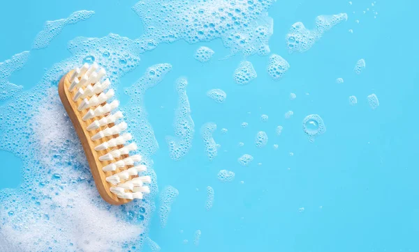 Wooden brush for cleaning clothes with foam of powder detergent water dissolution on blue backgrond. Top view