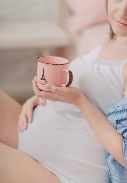 Barriga de uma mulher grávida, e em suas mãos uma caneca rosa — Fotografia de Stock