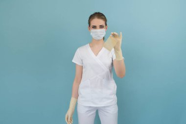 Doktorun elinde mavi arka planda eldiven ve maskeyle yanmış tıbbi bir yara bandı var.