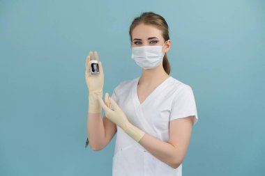 Maskeli ve eldivenli bir doktor mavi arka planda parmağında bir tonometre tutuyor.