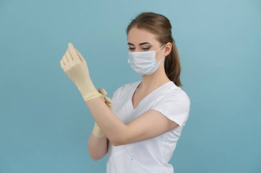Maskeli kadın doktor mavi arka planda lastik eldiven takıyor.