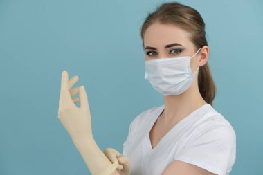 Maskeli kadın doktor mavi arka planda lastik eldiven takıyor.