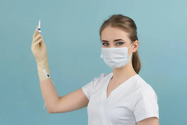 Maskeli ve eldivenli doktor mavi arka plandaki bir damlaya kateter takıyor.