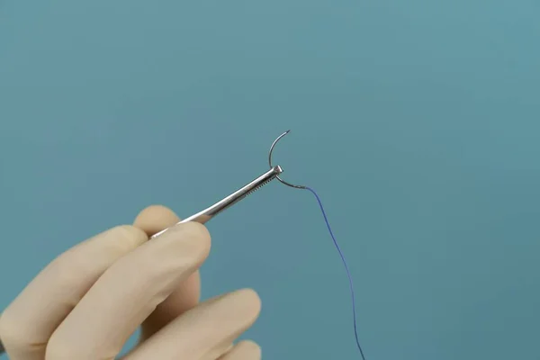 Doktor, mavi arka planda maske takan eldivenlere iğne batırılmış ameliyat ipliği tutuyor.