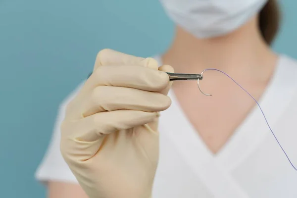 Doktor, mavi arka planda maske takan eldivenlere iğne batırılmış ameliyat ipliği tutuyor.