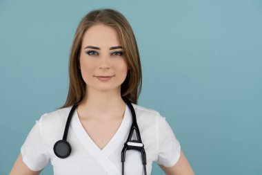 Beyaz kadın doktor kameraya bakıyor boynunda steteskopla mavi arka planda.