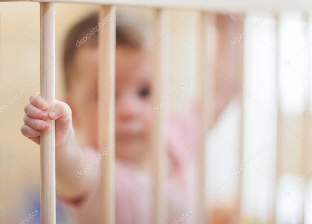 Sad baby stay alone in crib frame. Child tiny hands holding onto the side of the crib. Help for kids, adoption concept photo. Portrait of little lonely baby standing in crib. Waif child. Loneliness